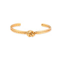 Gold cuff bracelet with twisted rope texture and decorative knot centerpiece on cream background.